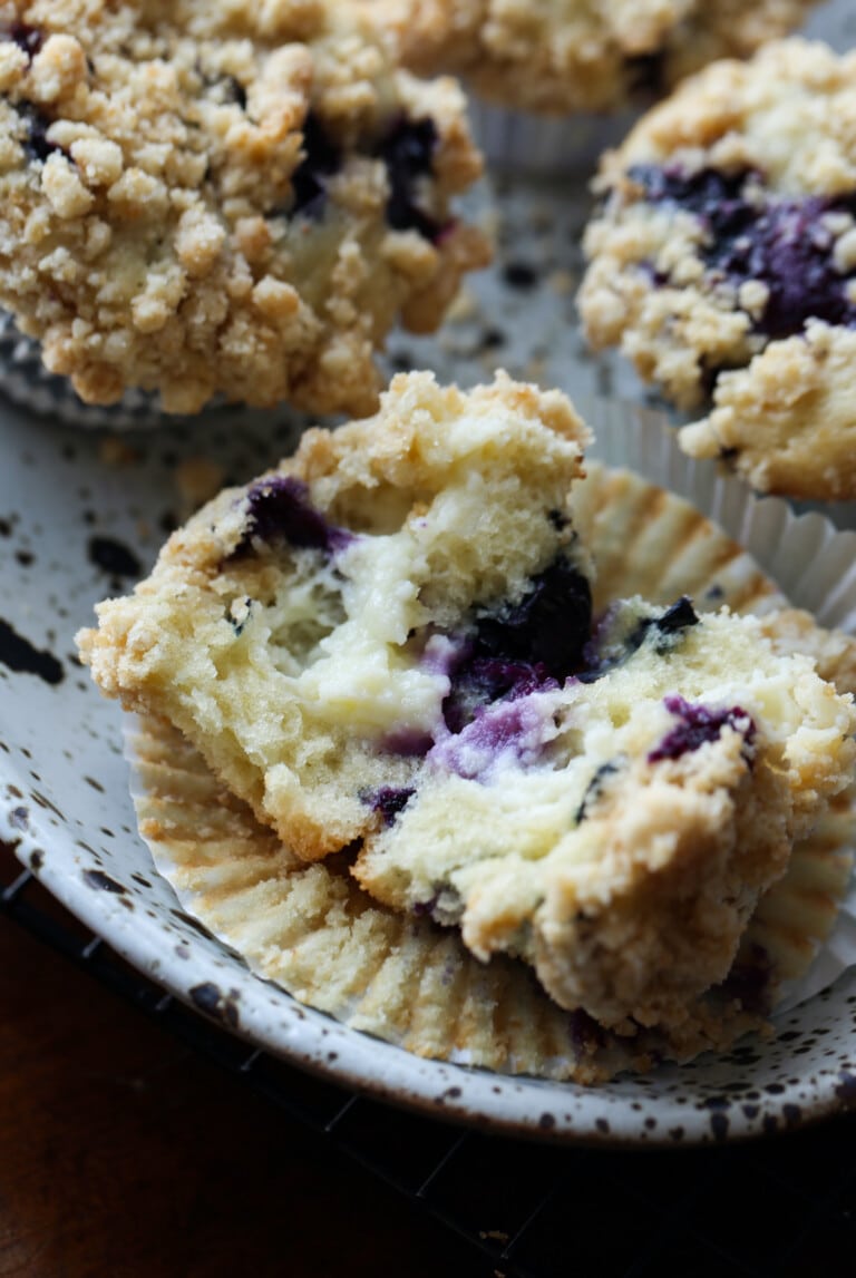 A blueberry muffin broken open to reveal the cream cheese filling, next to more muffins on a plate.