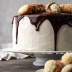 Cannoli cake on a cake stand drizzled with chocolate ganache and garnished with cannoli pastries.