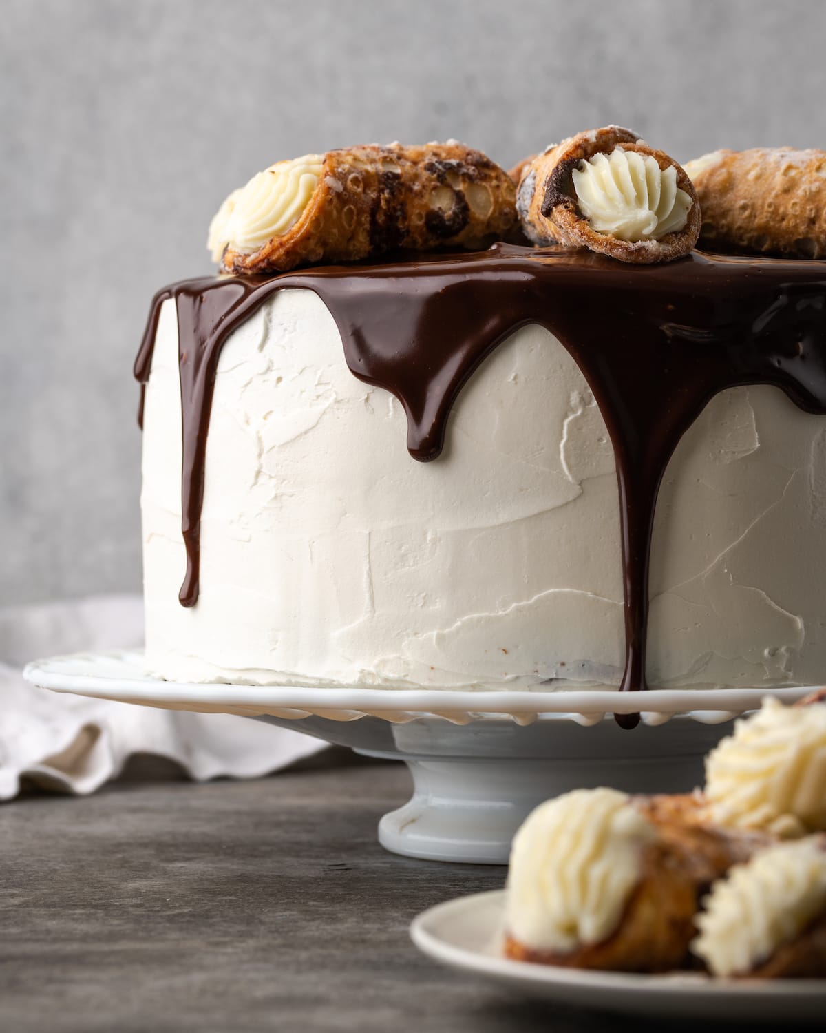 Cannoli cake on a cake stand drizzled with chocolate ganache and garnished with cannoli pastries.