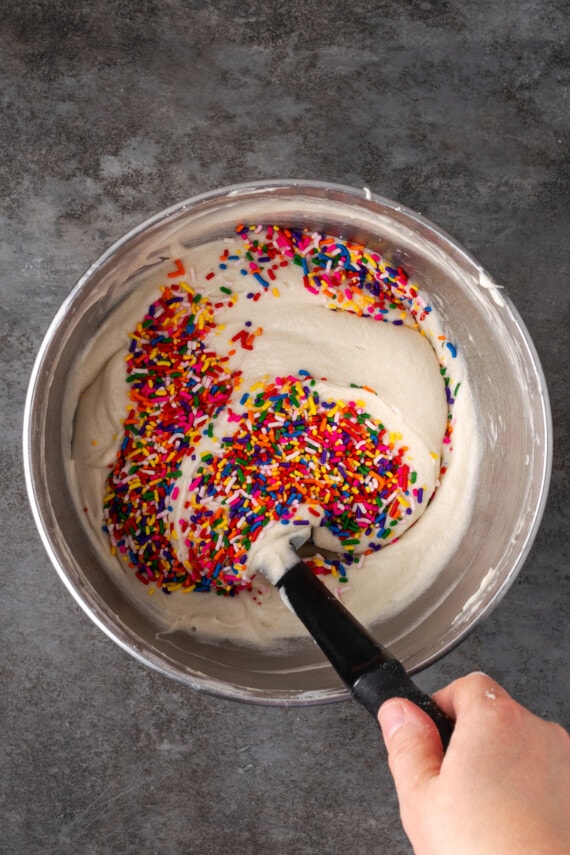 A hand using a black spoon to stir rainbow sprinkles into cake batter in a metal bowl.