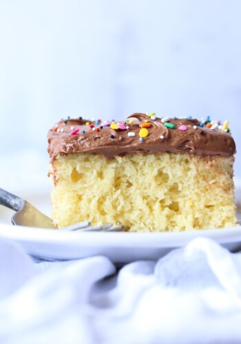 Vanilla cake with chocolate frosting and rainbow sprinkles on a white plate with a fork.
