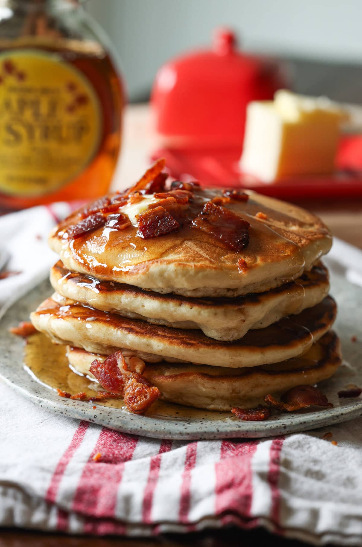 A stack of Maple Bacon Pancakes topped with butter, more chopped bacon, and maple syrup
