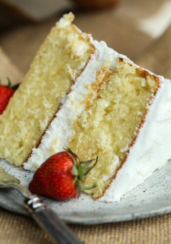 A slice of frosted vanilla cake on a plate next to a fresh strawberry.
