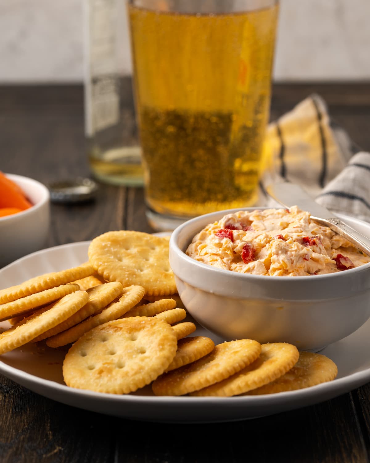A bowl of pimento cheese served on a platter of crackers, with glasses of beer in the background.