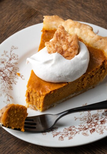 A slice of pumpkin pie on a plate topped with whipped cream and a sugar cookie, with a forkful of pie missing.