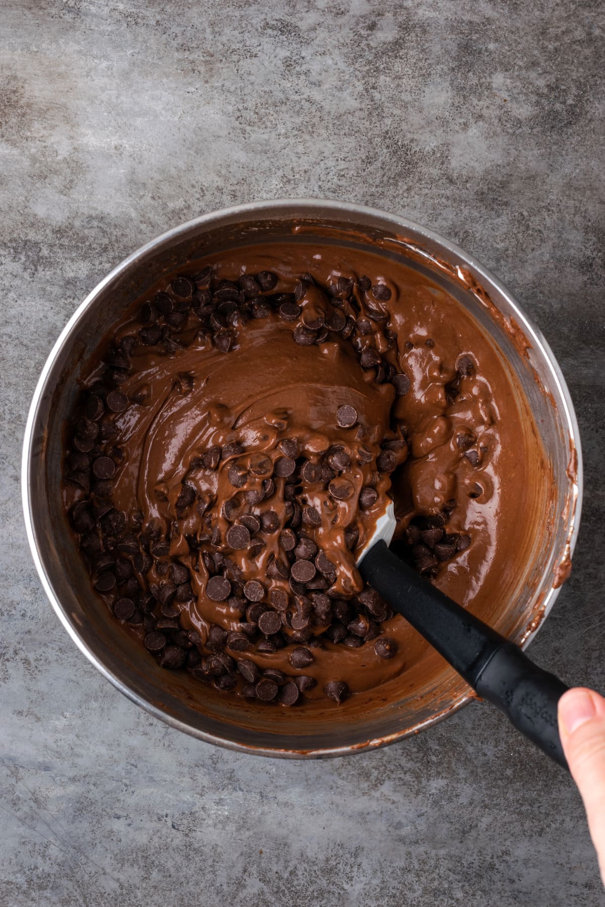 Chocolate chips are stirred into chocolate cake batter in a metal mixing bowl.