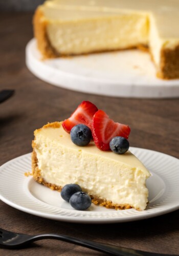 A slice of sour cream cheesecake garnished with fresh berries on a white plate, with the full cheesecake in the background.