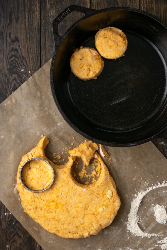 A round biscuit cutter is used to cut out sweet potato biscuit dough next to two biscuits in a cast iron skillet.