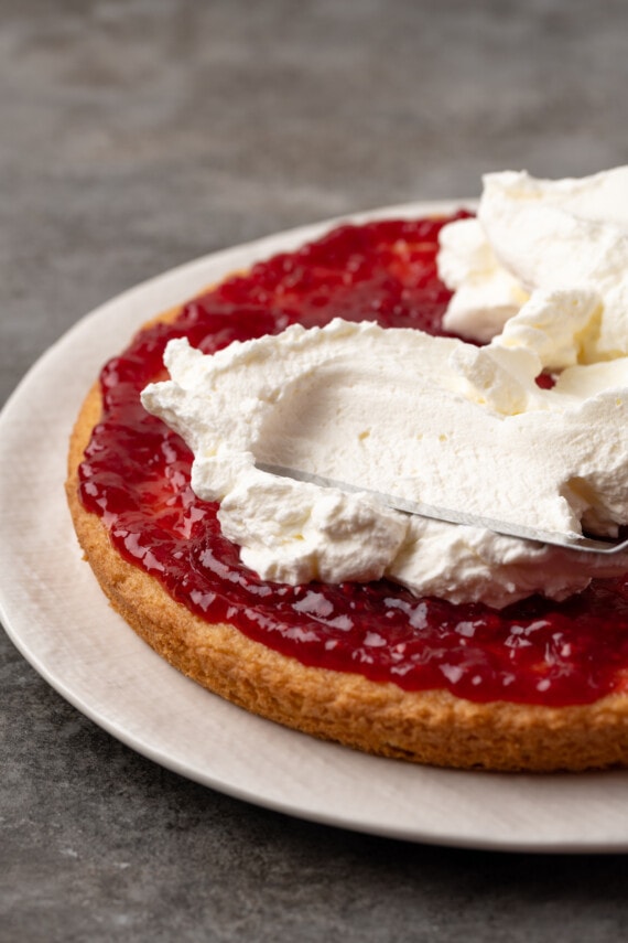 An offset spatula spreads whipped cream over the jam layer on a sponge cake.