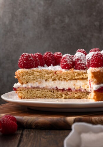 Victoria sponge cake topped with fresh raspberries on a white plate with a large slice missing.