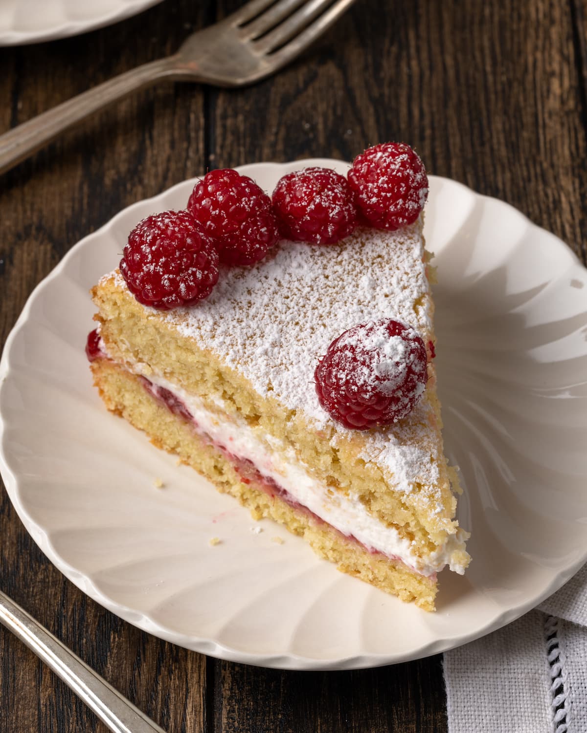 A slice of Victoria sponge cake on a white plate, topped with fresh raspberries.